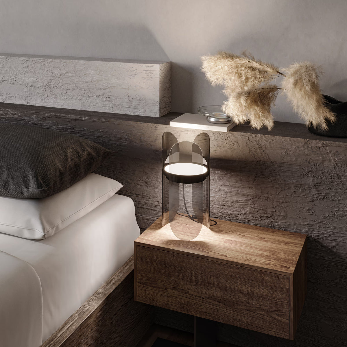 A modern table lamp with a clear curved glass shade and a white illuminated circular top, placed on a wooden bedside table in a bedroom setting next to a bed.